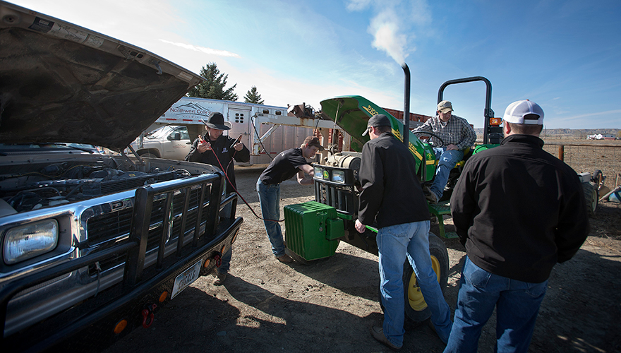 Group of male students outside in front of a horse trailer connecting a tractor to a pickup truck with jumper cables