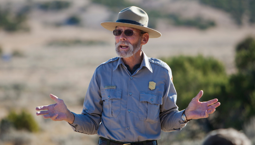 An older male park ranger with his arms spread talking to a crowd with a mountain, small trees, and desert landscape in the background