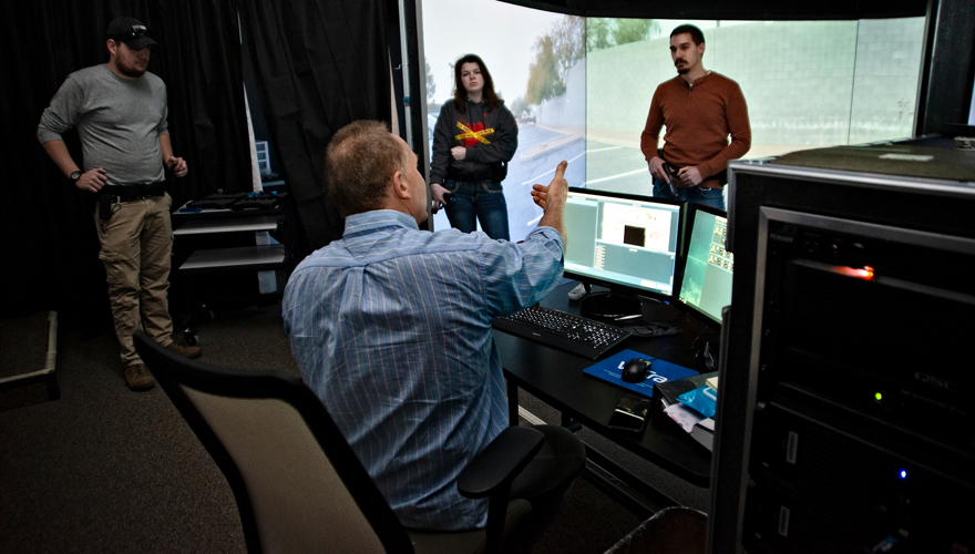 Male instructor talking to three students on one side of a 270 degree simulator screen sitting at a computer control system