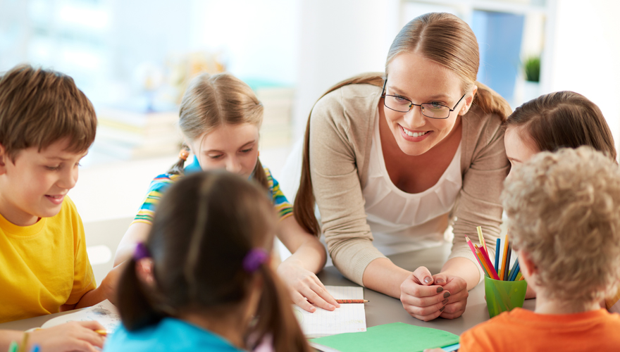 Female teacher leaning on a table with boys and girls doing an art project