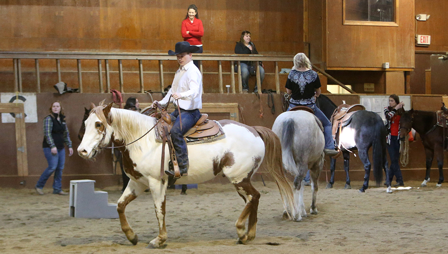 Man riding a white and brown horse in an indoor areans with other people and horses behind him and people watching from above