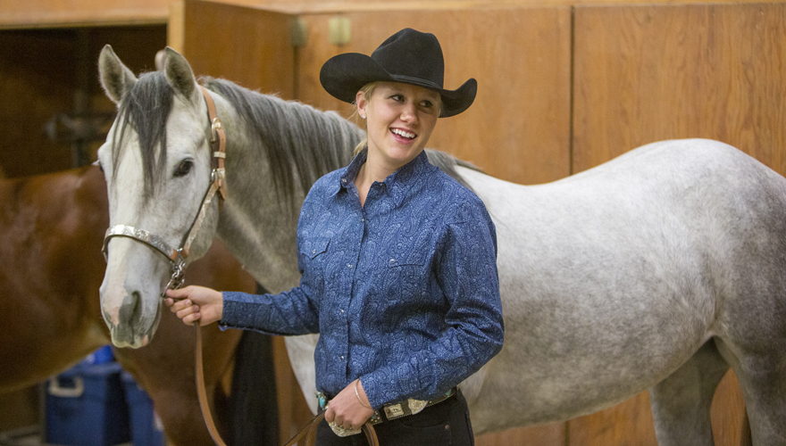 Smiling female student in a black hat holding the reins of a gray colored horse