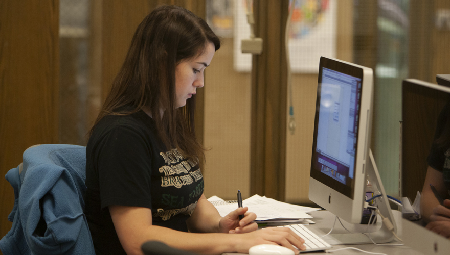 Female student sitting in front of a screen with one hand on a keyboard and the other holding a pen