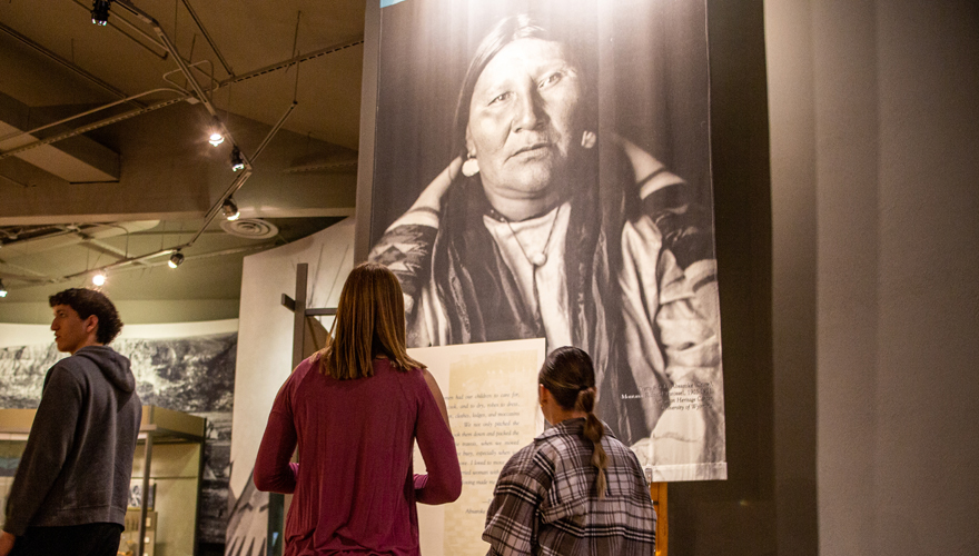 Two female students in a museum looking up at a large Native American portrait hanging from the ceiling