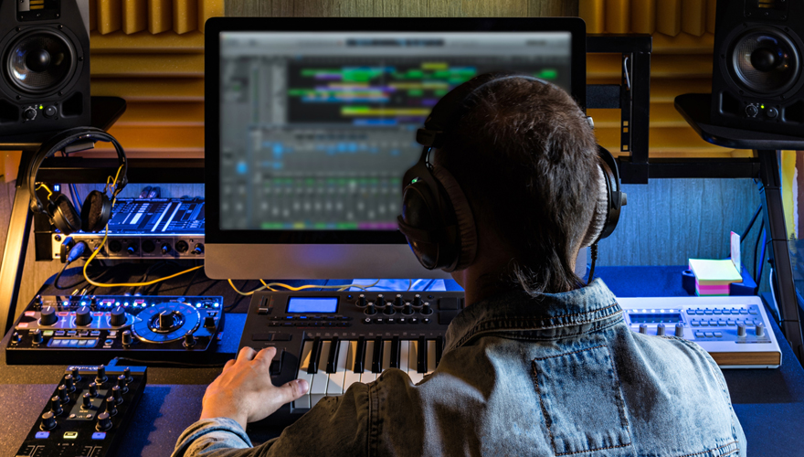 A male student wearing headphones sitting at a board with levers and switches with a computer screen and speakers in front of him