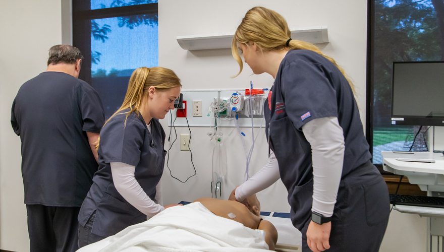 Two female nursing students tending to a mannequin in a hospital bed with an instructor looking at a monitor in the background next to a window