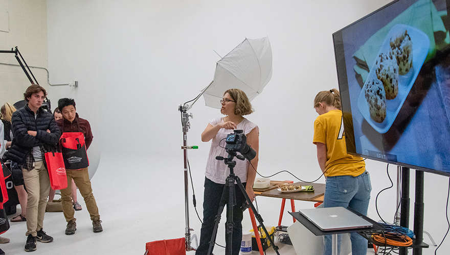 Photography professor pointing to a camera and taliing to students with large screen showing muffins behind her