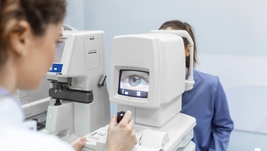 Female in white lab coast giving a patient an eye exam with image of an eye on a small screen in front of her