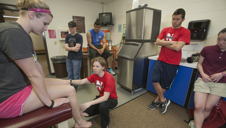 Crouching female instructor with students standing behind her in an athletic training room working on a female student's knee who is sitting on a bed