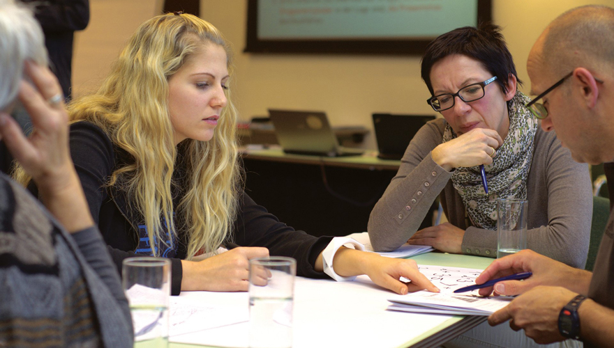 Adult students sitting around a table filled with paper and water glasses with two of them pointing to a notepad