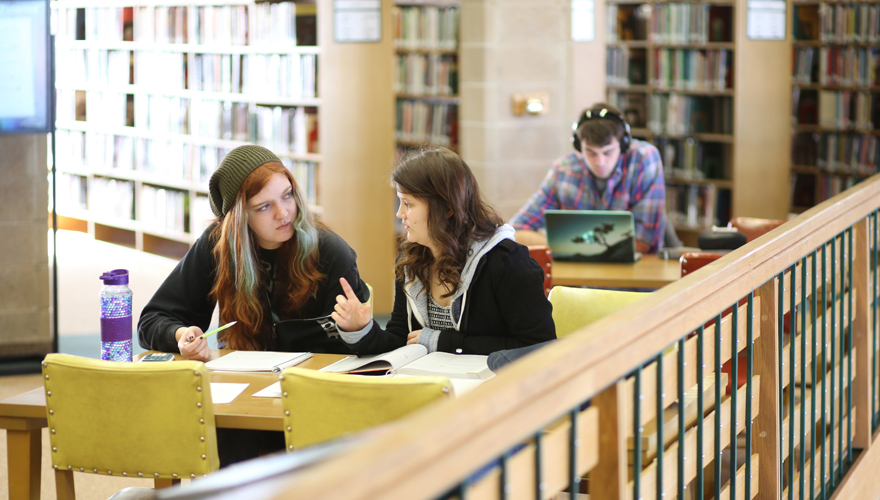 Two female students talking at a table with books and a water bottle on the second floor of Hinckley Library