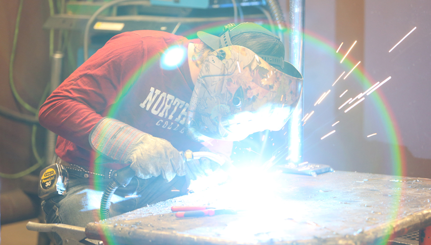 Male student in a red Northwest College shirt wearing a welding shield working at a table with a lens flare from bright sparks in front of him