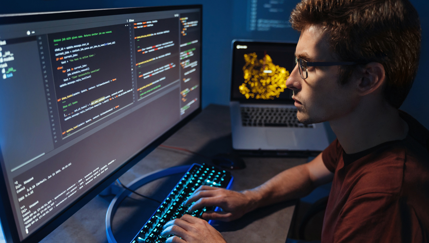 Male student wearing glasses sitting a computer console in a dimly lit room with code on the screen