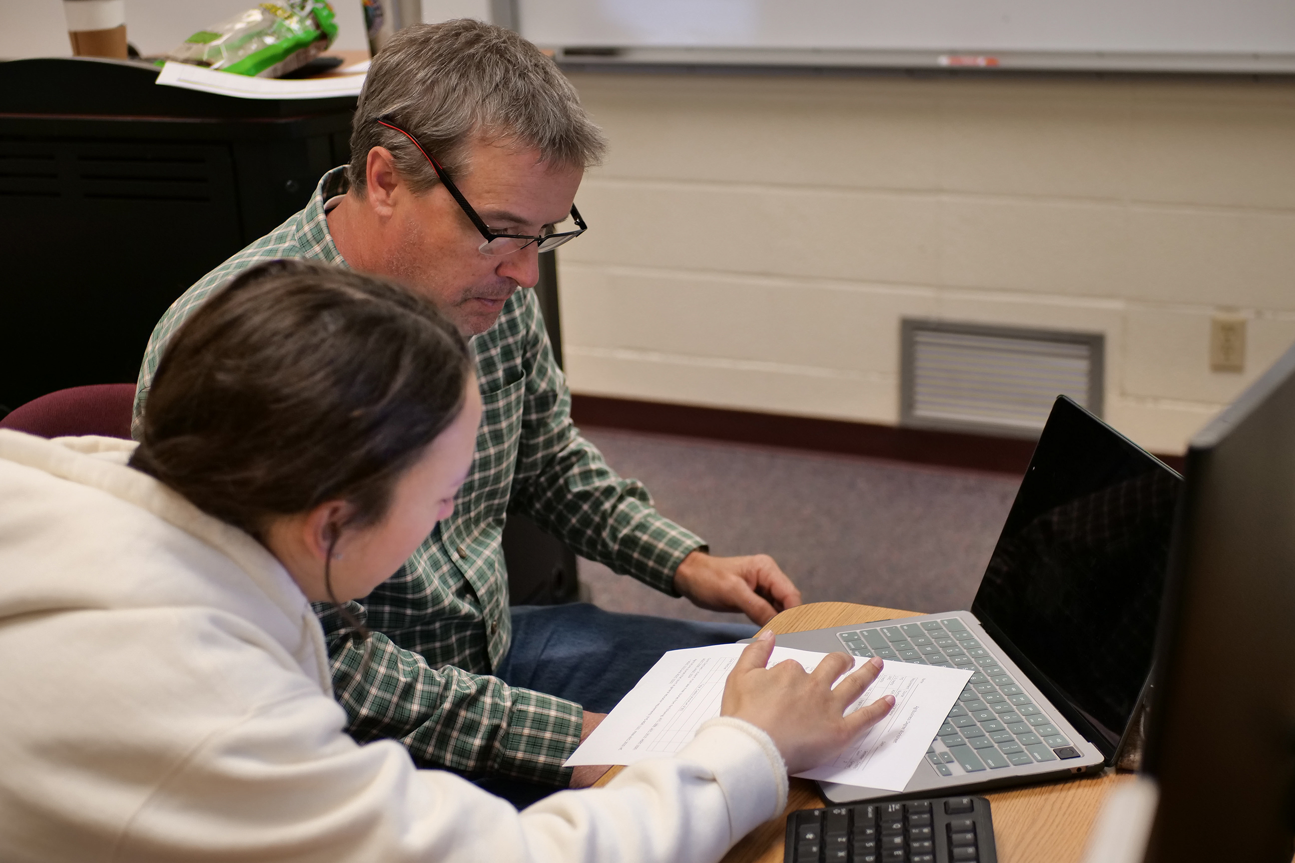 A professor sits with a student by a laptop with class schedule