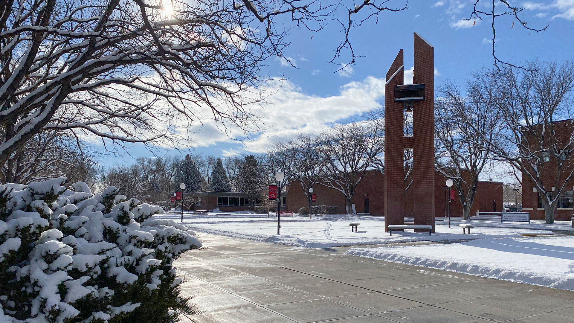 Campus mall in winter with carillon set against a blue sky with scattered clouds, snow-covered bushes, snow on the ground and dry sidewalks