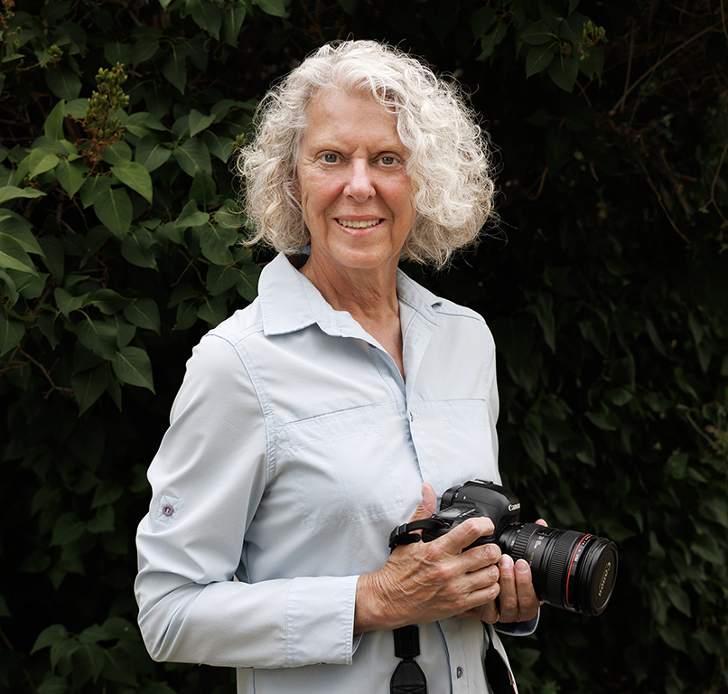 A blond-haired woman in a light blue shirt holding a camera in front of green leaves on a tree