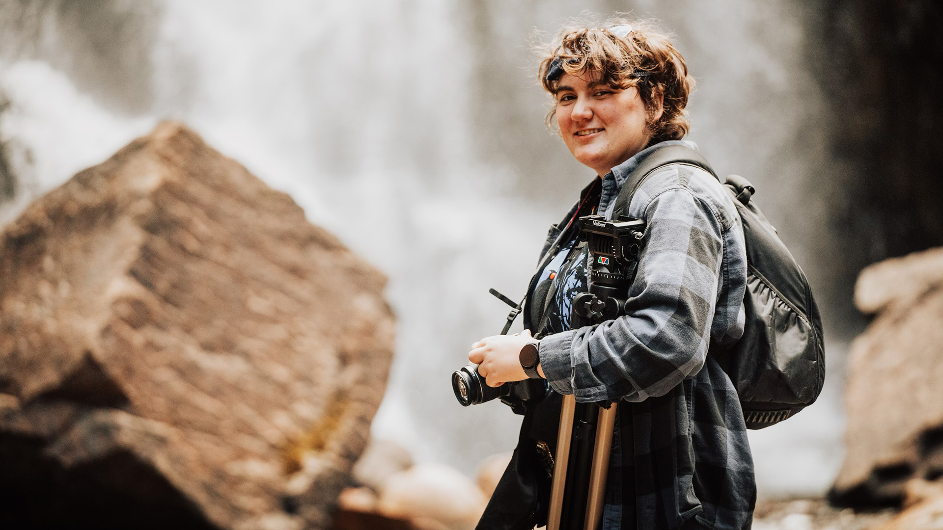 A female student holding a camera and tripod in front of boulders and a waterfall