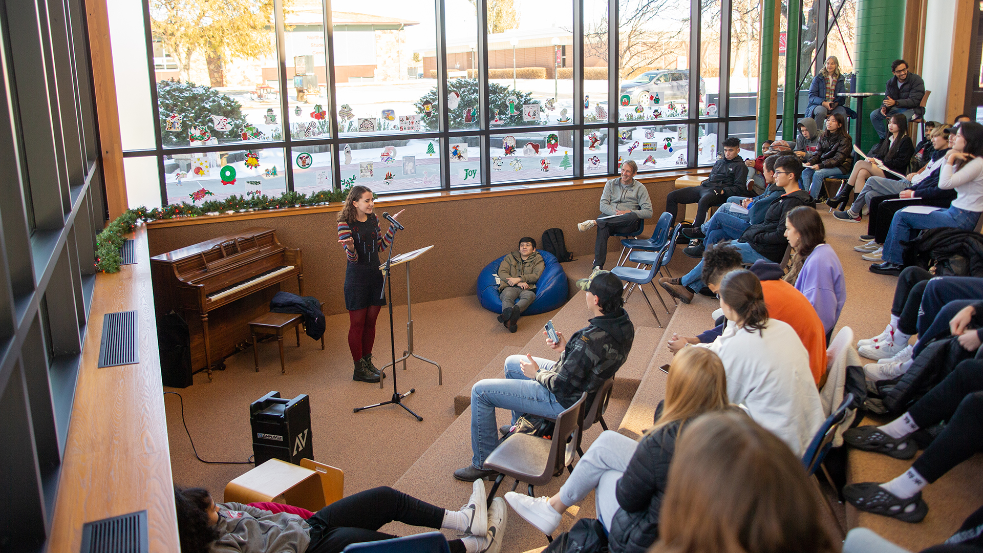 Female student with arms spread in front of a microphone and podium talking to a group of people in library amphitheater