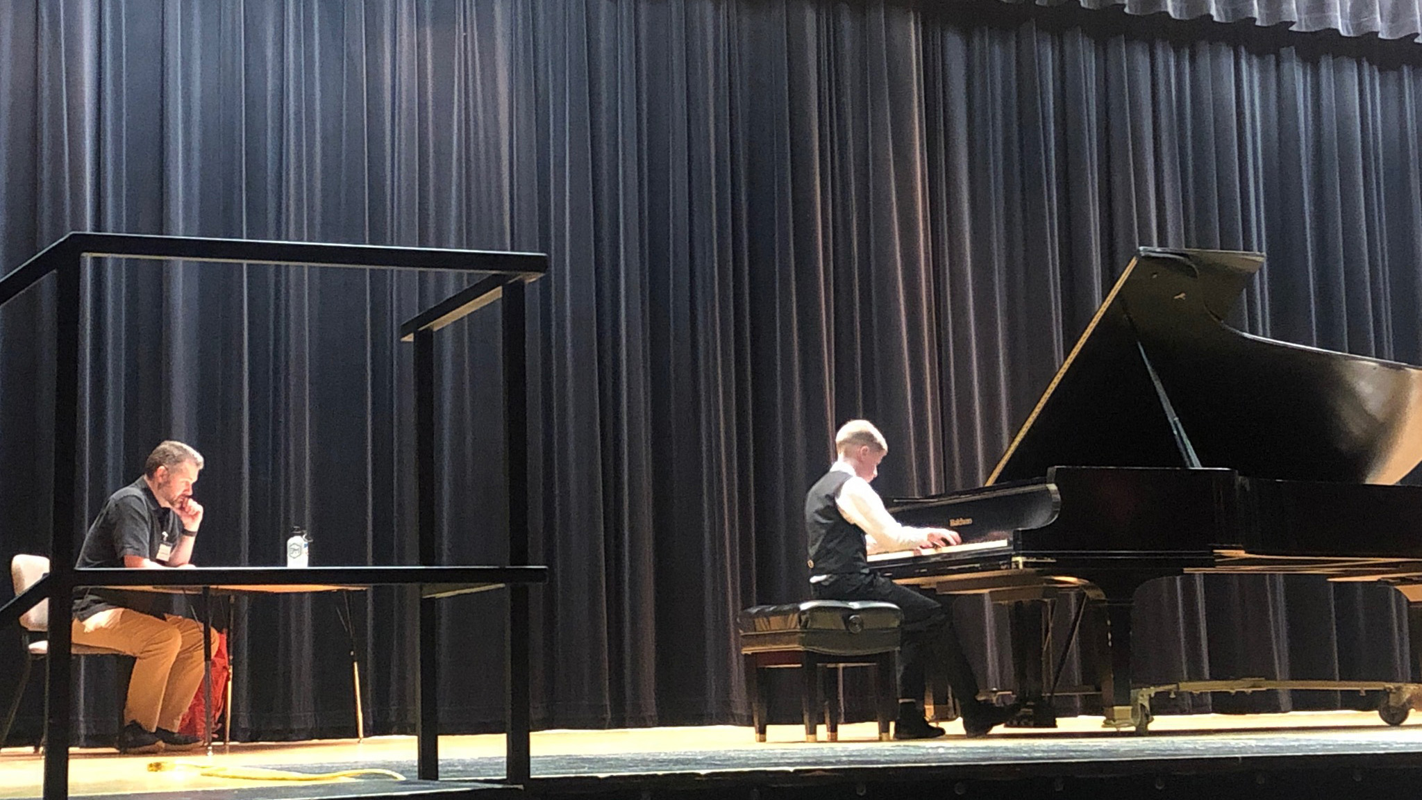 A boy playing a piano on stage with a man sitting at a table behind him in front of dark curtains