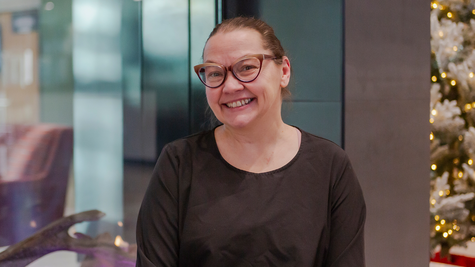 Woman in black shirt wearing glasses sitting in front of a fireplace with glass front and a Christmas tree in the background